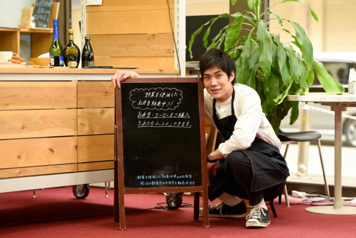 自店舗の看板前に座っている笑顔の経営者