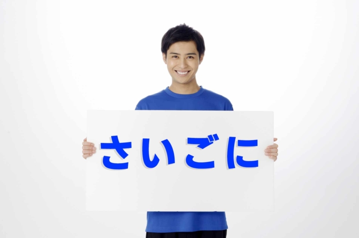 「さいごに」と書かれたプラカードを胸の前に持ち、笑顔で立っている男性