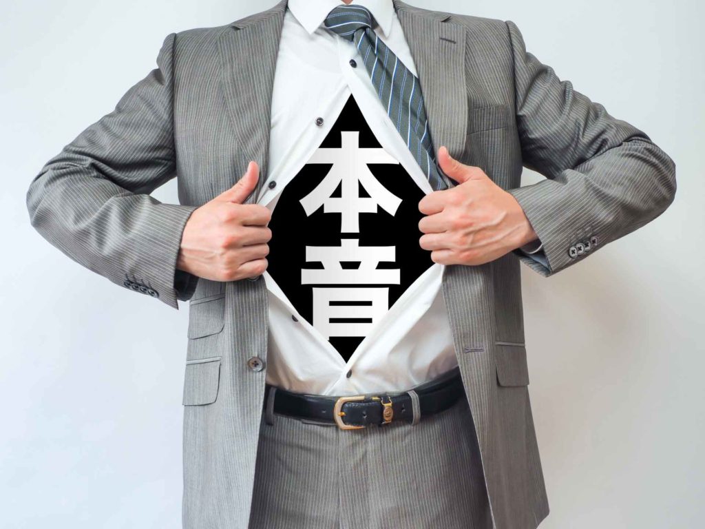 シャツを開いた部分に「本音」と書かれたスーツ姿の男性