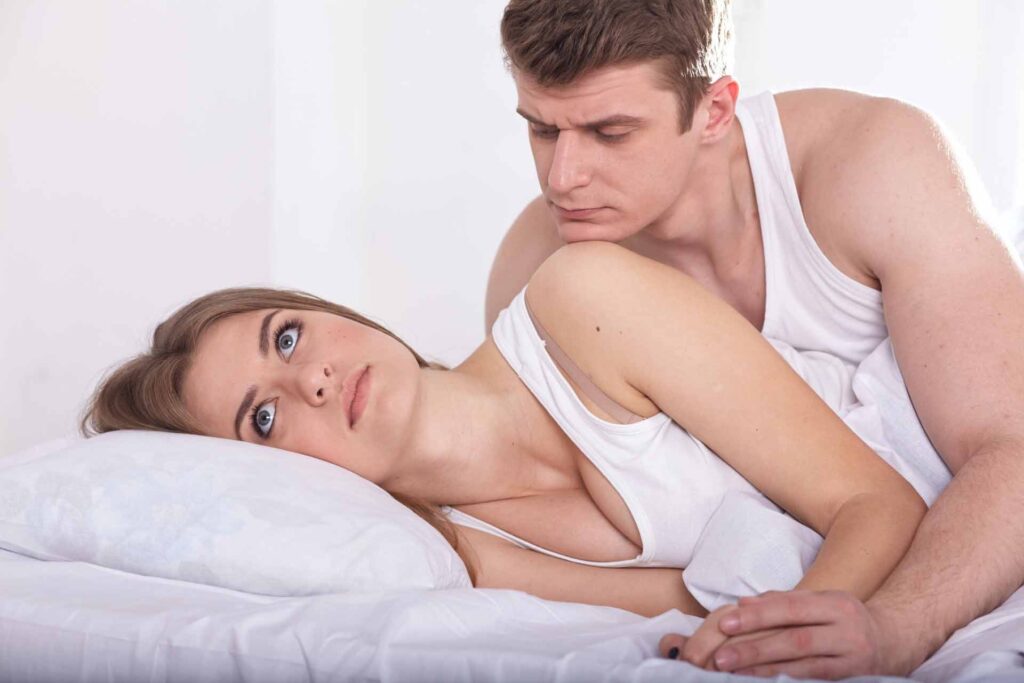 ベッドで機嫌悪そうにしている女性と、女性の顔を覗き込みながら話している男性