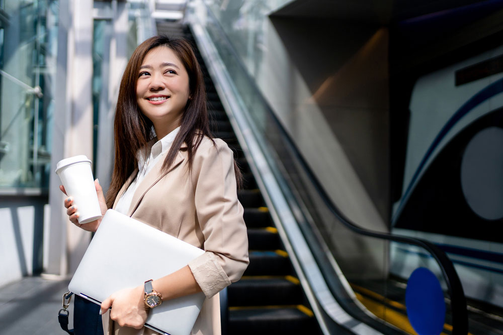 エスカレーターの前でノートPCと珈琲を手に持つ女性